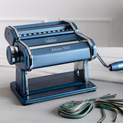 Marcato | Atlas 150 Pasta Maker (Ice) on a table, showcasing its manual handle and adjustable dial for rolling and cutting fresh pasta dough.
