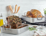 Hestan Provisions Classic Roaster with Rack on stovetop, featuring roast turkey and beef, surrounded by kitchen utensils, olive oil, herbs, and a towel.