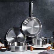 Heritage Steel | Eater 5 Piece Essentials Set: stainless-steel cookware including sauté pan, skillet, and saucepan, artfully arranged with garlic, salt, and olive oil on a kitchen countertop.