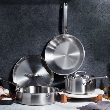 Heritage Steel | Eater 5 Piece Essentials Set: stainless-steel cookware including sauté pan, skillet, and saucepan, artfully arranged with garlic, salt, and olive oil on a kitchen countertop.