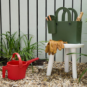 Hachiman | Garden Tool Bag Garden Green on a white stool, holding gardening tools, beside a red scooping container on gravel, ideal for organized garden work.