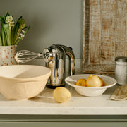 Dualit Hand Mixer in polished chrome with batter-covered whisk, set with a ceramic mixing bowl and lemons, suggesting a lemon baking project.