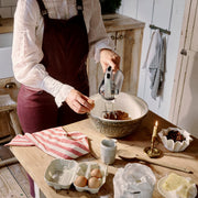 A woman uses a Dualit Hand Mixer, Polished Chrome, to blend ingredients in a rustic kitchen, surrounded by baking essentials like eggs, flour, and butter.