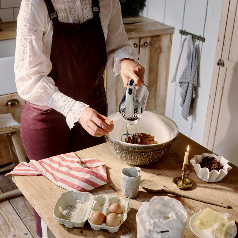 A woman uses a Dualit Hand Mixer, Polished Chrome, to blend ingredients in a rustic kitchen, surrounded by baking essentials like eggs, flour, and butter.