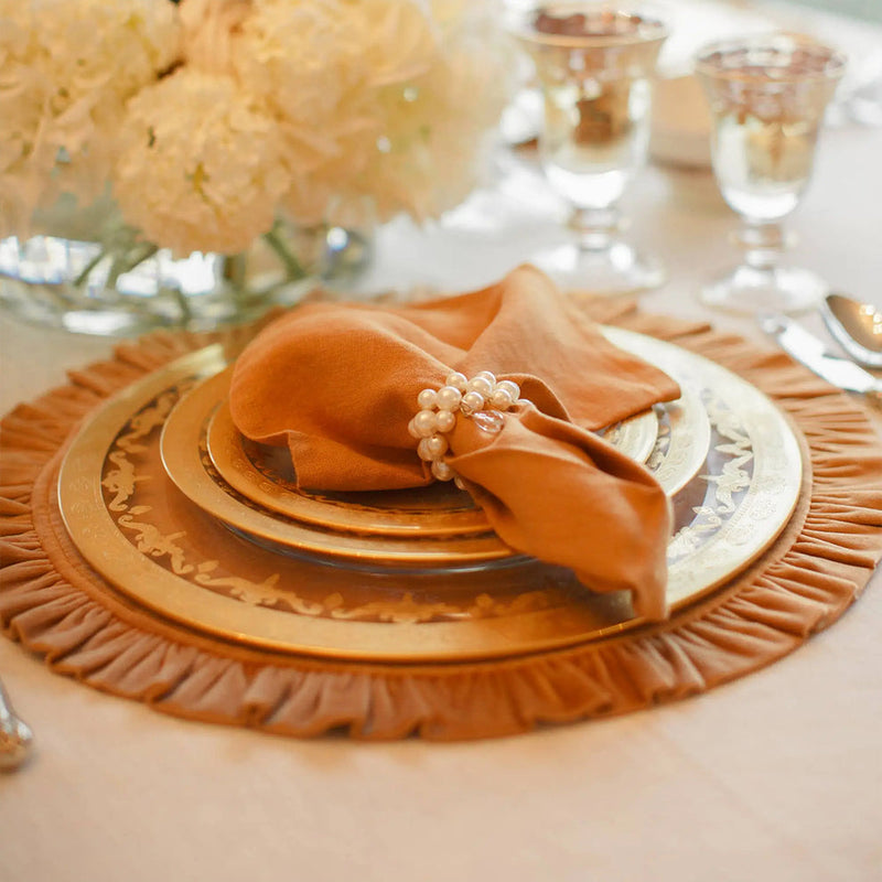 Formal dining setup featuring the Arte Italica & Crown Linen Designs Round Ruffle Placemat, highlighting its elegant gold accents, under a stack of dinnerware and a napkin.