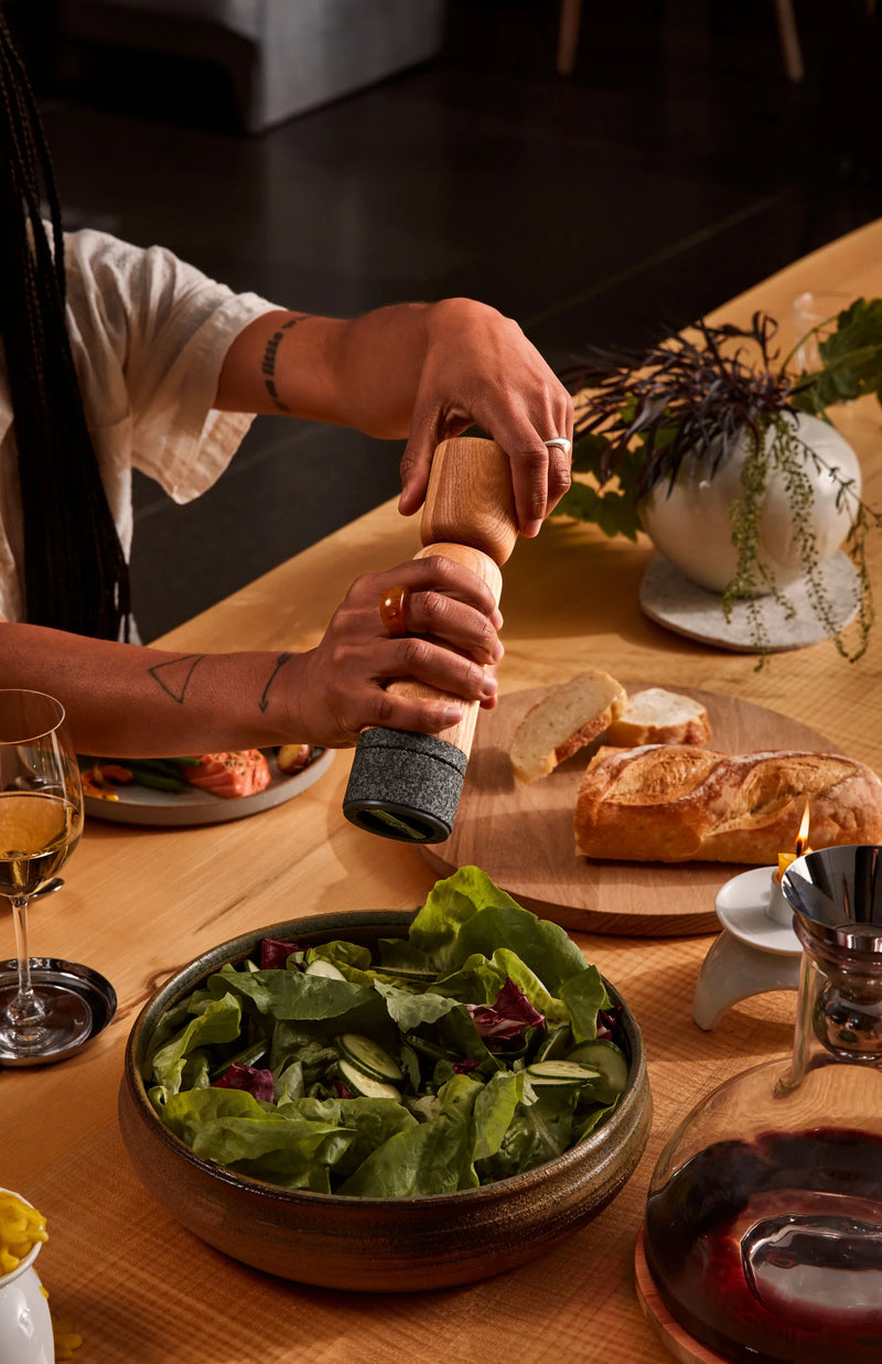 Holcomb Studio | Stone Pepper Mill in use, held over a dining table with salad, wine, and bread, highlighting its elegant design and functionality.