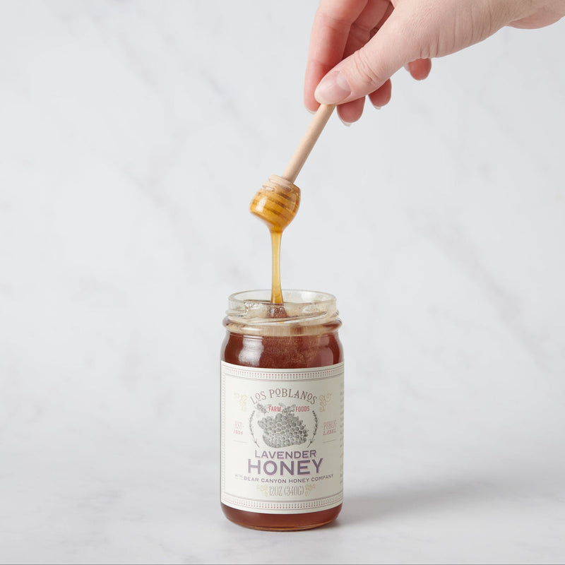 Los Poblanos Lavender Honey jar with a wooden dipper, being held above the jar, highlighting the specialty honey product available at Athens Cooks.