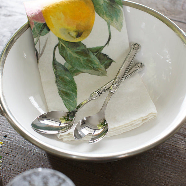 Arte Italica & Crown Linen Designs Hotel Collection Salad Servers rest elegantly on a cloth napkin in a white enamel bowl, showcasing vintage elegance and craftsmanship.