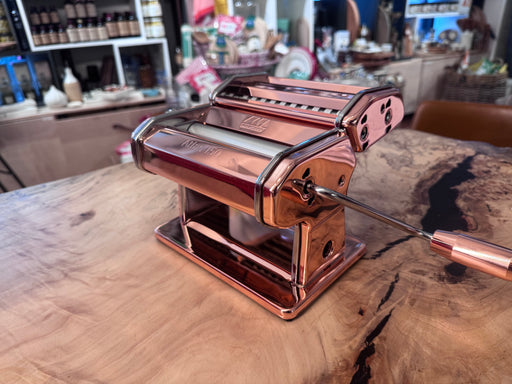 Marcato | Atlas 150 Pasta Machine (Copper) on a wooden countertop, featuring a crank handle for crafting diverse pasta shapes, showcasing durability and precision perfect for home chefs.