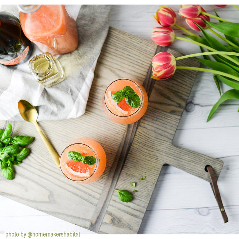 J.K. Adams | 1761 Ash Driftwood Square Serving Board elegantly displays two pink grapefruit drinks with basil, accompanied by a carafe, jar, and gold-tone spoon.