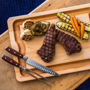 J.K. Adams | Maple BBQ Carving Boards, No Personalization Medium, topped with grilled steak, roasted garlic, artichoke, zucchini, and carrots, framed by a carving knife and fork.