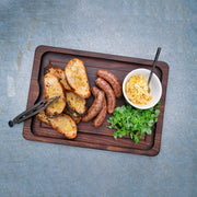 J.K. Adams Walnut BBQ Carving Board displayed with grilled sausages, crostini, shredded cheese, herbs, and tongs, highlighting its versatility for BBQ and family meals.