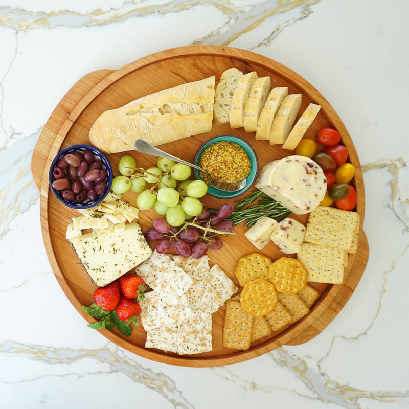 J.K. Adams Cherry Round Wooden Serving Tray artfully arranged with cheeses, baguette slices, olives, fruits, and more, showcasing its elegance and functionality for entertaining.