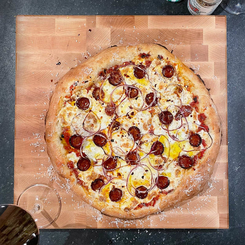 J.K. Adams Professional End Grain Board in maple, showcasing a freshly-baked pizza with mozzarella, pepperoni, red onion, and chili flakes, next to wine and seasoning.