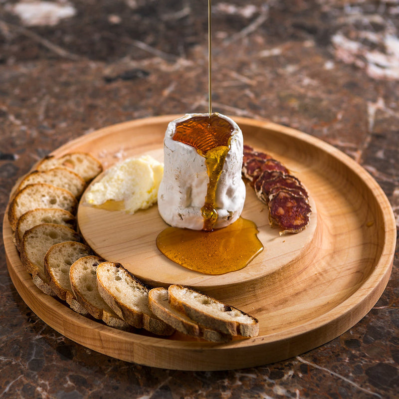 J.K. Adams Maple Round Cheese Board with Cracker Groove, 12 Inches, showcasing cheese, charcuterie, and honey drizzle, with baguette slices around the edge.