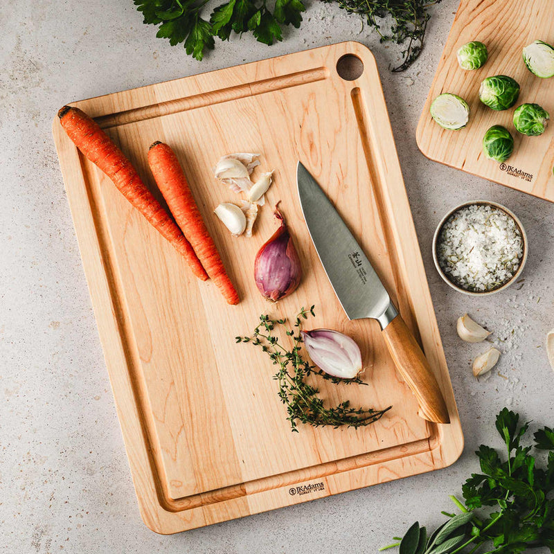 J.K. Adams Maple Prep Cutting Board with Juice Groove featuring fresh vegetables, a kitchen knife, and a bowl of rosemary salt, ideal for culinary preparation.