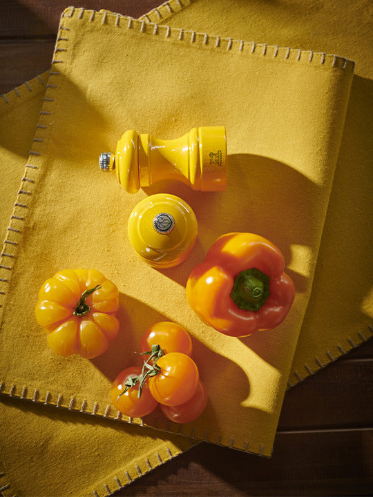 Peugeot | Bistro Salt Mill, Saffron, showcased on mustard fabric with yellow tomatoes and bell pepper, highlighting its craftsmanship and color.