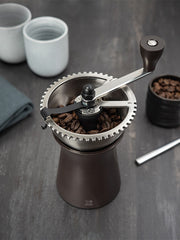 Peugeot | Kronos Coffee Mill, 7.5 inches, with whole beans in hopper and hand-crank, surrounded by ceramic cups and extra beans on dark surface.