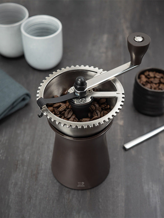 Peugeot | Kronos Coffee Mill, 7.5 inches, with whole beans in hopper and hand-crank, surrounded by ceramic cups and extra beans on dark surface.