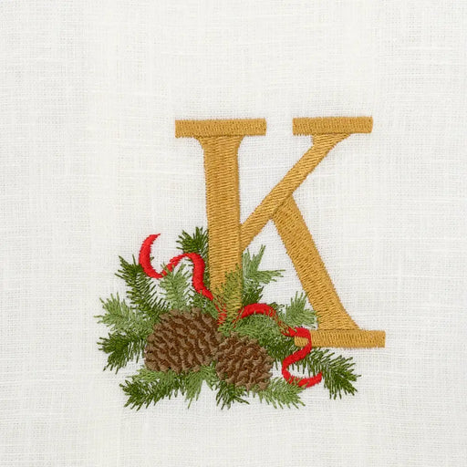 Arte Italica & Crown Linen Designs Winter Pine Monogram Towel features a golden K with festive sprigs, pine cones, and ribbon on white linen.
