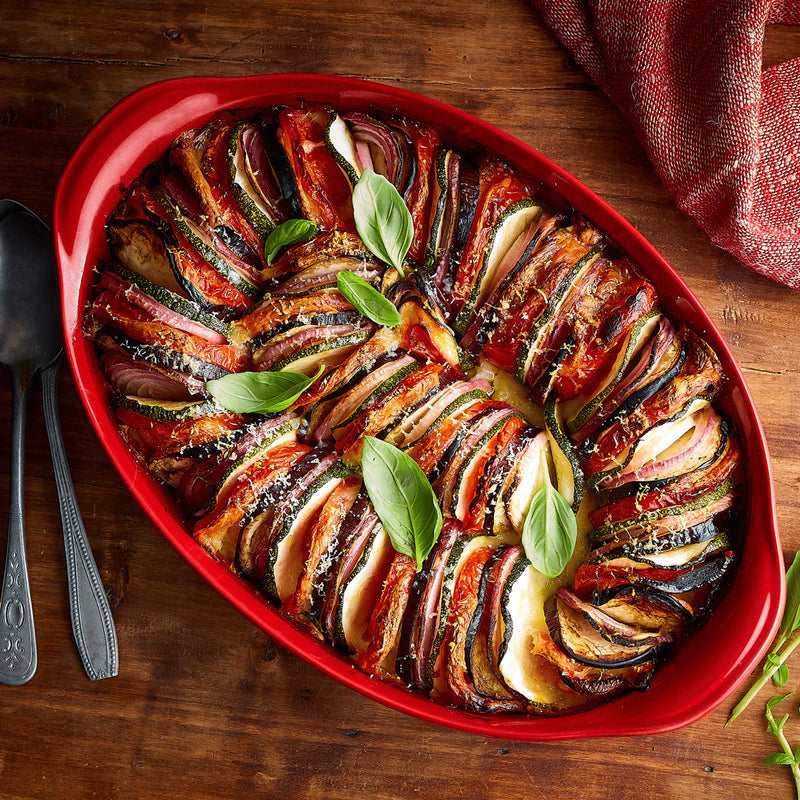 Emile Henry Large Oval Dish in red, showcasing a layered vegetable casserole with zucchini, eggplant, tomato, and onion, perfectly complementing Athens Cooks' curated kitchenware.
