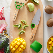 Opinel | Le Petit Chef 3pc Knife Set, Green displayed with a paring knife, Y-peeler, silicone sheath, and fresh fruit on a wooden cutting board.