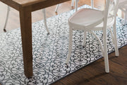 Liberty L Italian Kitchen Sink Floor Mat by Etú HOME, featuring a black and white geometric tile pattern, under a wooden chair and table leg on dark-wood flooring.