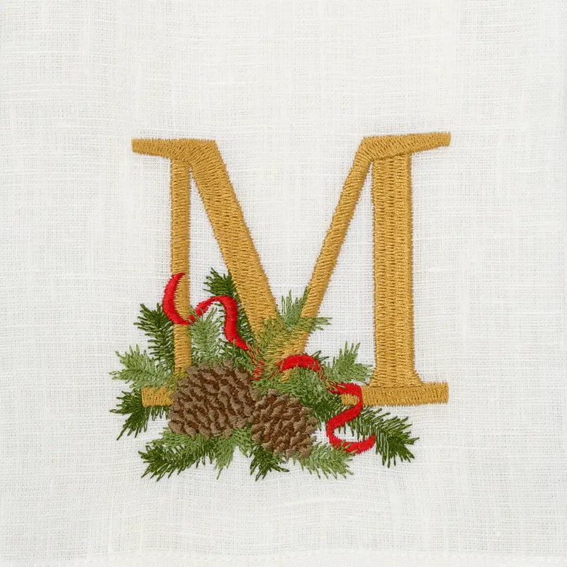 Winter Pine Monogram Towel, M, by Arte Italica & Crown Linen Designs, features an embroidered gold M with pine boughs, pinecones, and a red ribbon on white linen.