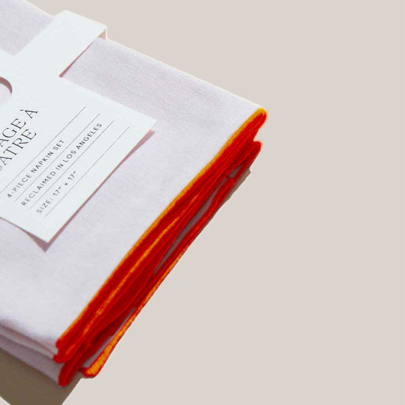 Atelier Saucier | Blush Linen Orange Napkins, Set of 4, features white napkins with orange trim, neatly bundled with a paper label, ideal for elegant tablescapes.