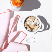 Atelier Saucier's Blush Linen Orange Napkins enhance a minimalist breakfast scene with shredded-wheat cereal, a white dish, and an orange ranunculus flower.