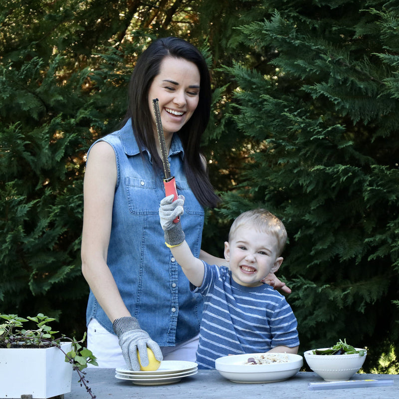Woman and boy in garden using Microplane Cut Resistant Kitchen Safety Gloves for Kids while preparing food at a stone table with dishes and herbs.