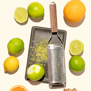Microplane | Master Series Fine Grater with Walnut Handle on baking sheet with grated citrus, surrounded by limes, lemon, orange slice, and ginger.