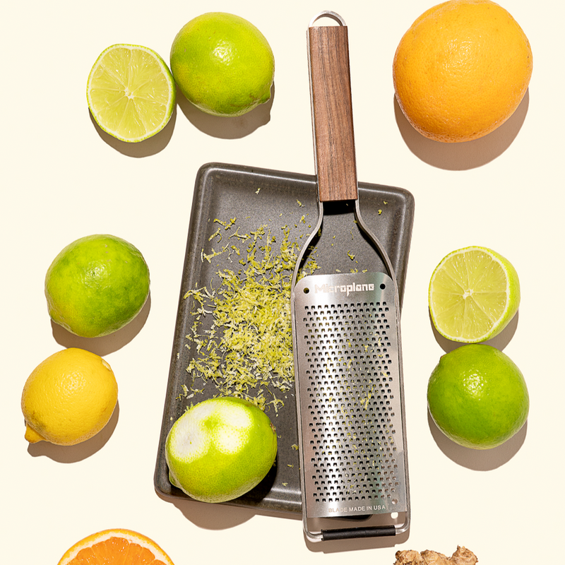 Microplane | Master Series Fine Grater with Walnut Handle on baking sheet with grated citrus, surrounded by limes, lemon, orange slice, and ginger.