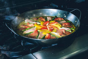 Matfer Bourgeat black carbon steel paella pan cooking vibrant paella with saffron rice, asparagus, tomatoes, peppers, chorizo, and meat strips on a gas burner.