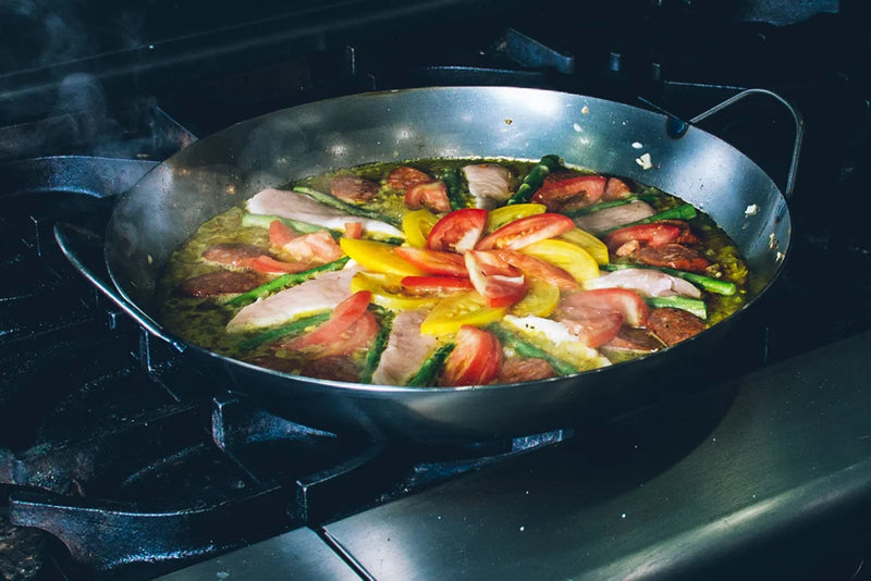 Matfer Bourgeat black carbon steel paella pan cooking vibrant paella with saffron rice, asparagus, tomatoes, peppers, chorizo, and meat strips on a gas burner.