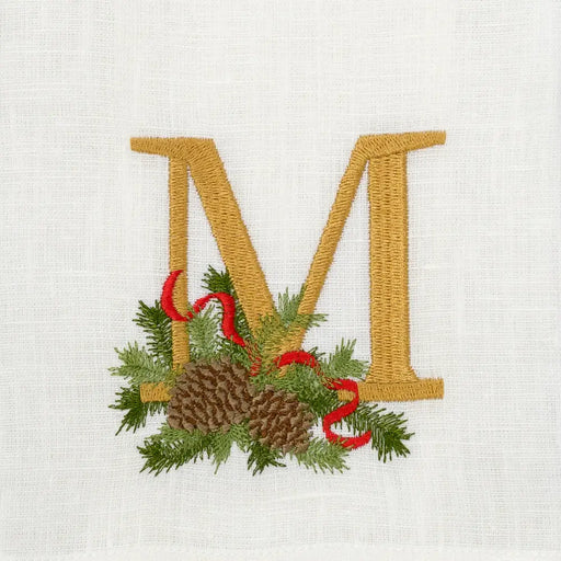 Winter Pine Monogram Towel, M, by Arte Italica & Crown Linen Designs, features an embroidered gold M with pine boughs, pinecones, and a red ribbon on white linen.