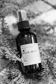 Among the Flowers | Man Mist, Wood Pile cologne bottle on pine branches, featuring cedarwood and sandalwood scents, embodying a rugged, natural aroma.
