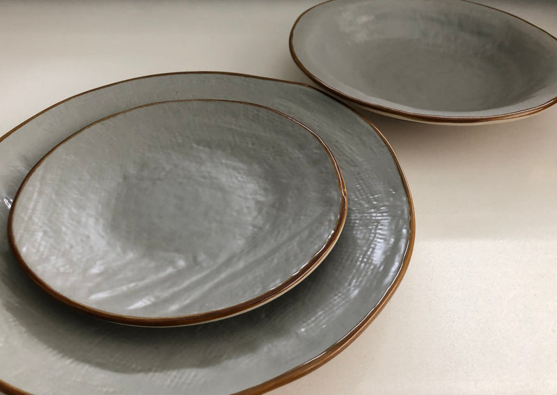 Close-up of Novita Mediterraneo stoneware dinner plates, showcasing unique hand-dipped craftsmanship in gray, highlighting their varied shapes and finishes.