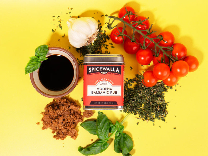 Spicewalla | Modena Balsamic Rub tin surrounded by garlic, cherry tomatoes, dried herbs, brown sugar, balsamic vinegar, and basil, suggesting a flavorful cooking preparation.