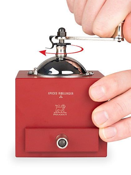 Peugeot | Roellinger Pepper Mill in red with a hand turning the metal crank, featuring a pull-out drawer and the “Épices Roellinger” label above the Peugeot logo.