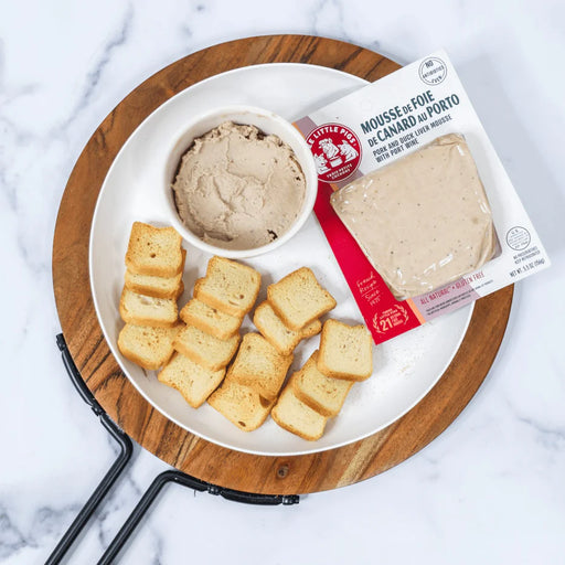 Three Little Pigs Duck Liver and Pork Mousse Pâté with Port Wine on a wooden board, served with crostini and its original packaging.