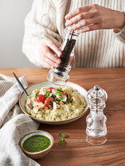 A hand in a cream sweater uses the Peugeot | Paris Nancy Pepper Mill, Acrylic, 9 inches, to season risotto on a cozy table setting.