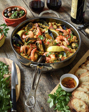 Hestan | NanoBond Cookware, 5qt Essential Pan + Lid, showcases seafood paella with shrimp, mussels, and herbs, surrounded by olives, wine, and bread on a rustic table.