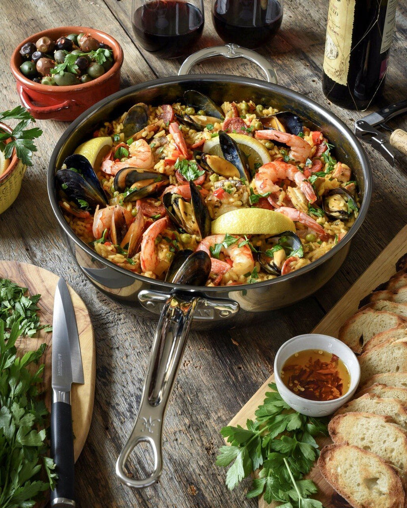 Hestan | NanoBond Cookware, 5qt Essential Pan + Lid, showcases seafood paella with shrimp, mussels, and herbs, surrounded by olives, wine, and bread on a rustic table.