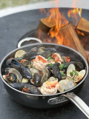 Hestan | NanoBond Cookware, 5qt Essential Pan + Lid, showcases a shellfish stew cooking over a wood fire, highlighting its versatility and durable titanium surface.