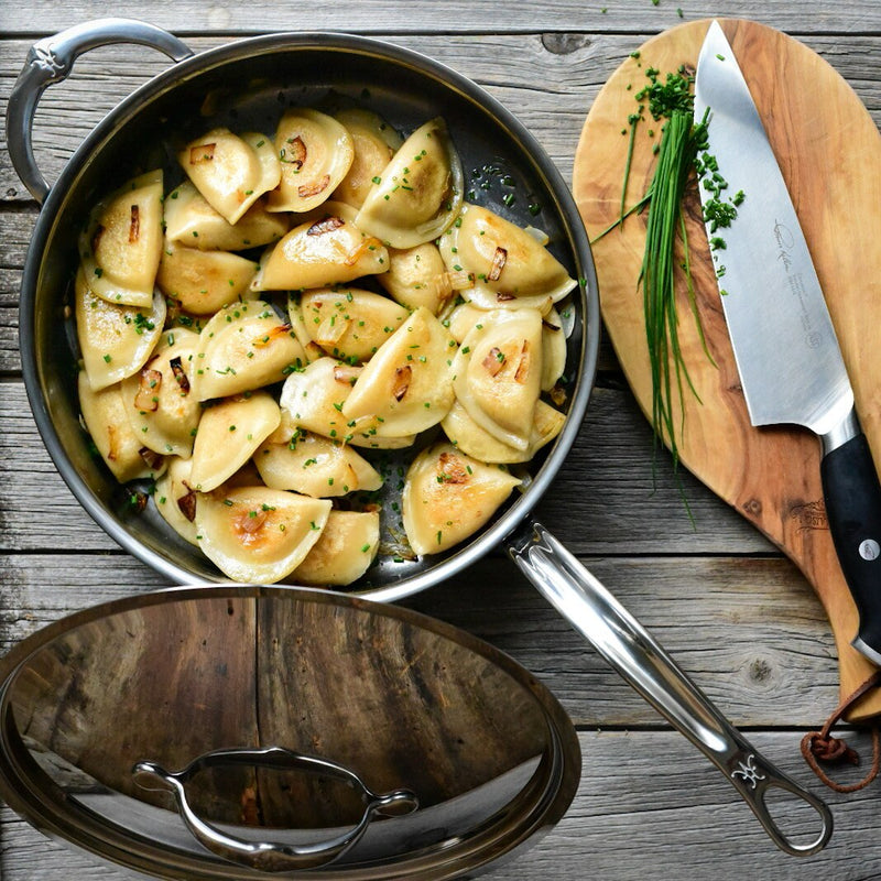 Hestan NanoBond 5qt Sauté Pan with caramelized onion pierogi, showcasing its versatile, durable design on a rustic wooden tabletop with fresh chives and a knife nearby.