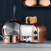 Dualit | 2 Slice Classic Toaster, Copper, on a marble countertop with matching copper cookware, showcasing its quality craftsmanship and unique design.