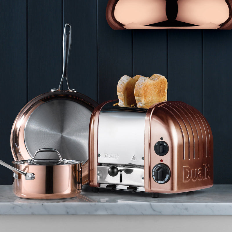 Dualit | 2 Slice Classic Toaster, Copper, on a marble countertop with matching copper cookware, showcasing its quality craftsmanship and unique design.