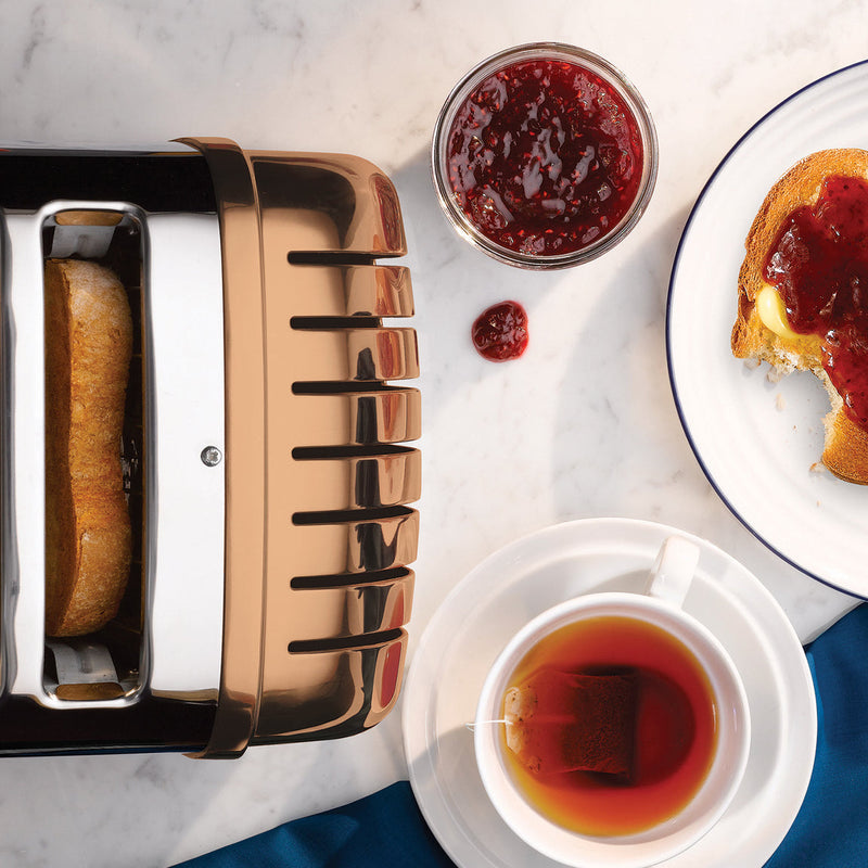 Dualit 2 Slice Classic Toaster, Copper, with toast popping up, accompanied by buttered toast with jam, tea, and jam jar in a cozy breakfast setting.
