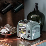 Dualit 2 Slice Classic Toaster, Eucalyptus, on a kitchen counter with rustic bread and vintage decor, showcasing its elegant design and craftsmanship.
